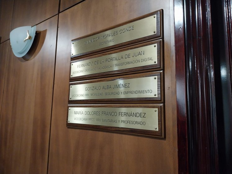 Rotulo en lat&oacute;n grabado en bajo relieve y patina de tinta negra, sobre metopa o peana de madera. Facultad de Medicina de la Universidad de Sevilla.