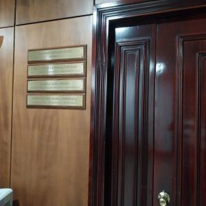 Rotulo en lat&oacute;n grabado en bajo relieve y patina de tinta negra, sobre metopa o peana de madera. Facultad de Medicina de la Universidad de Sevilla.